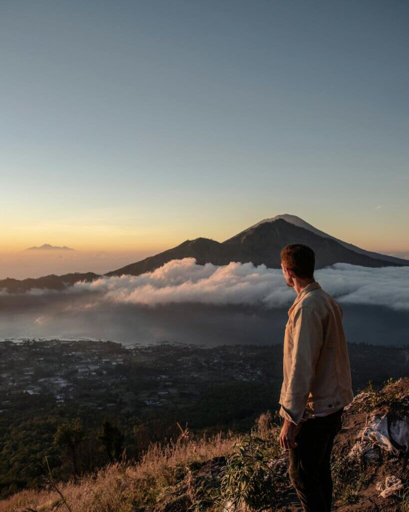 A man stands on a hillside overlooking Mount Batur amidst clouds during sunrise in Bali.