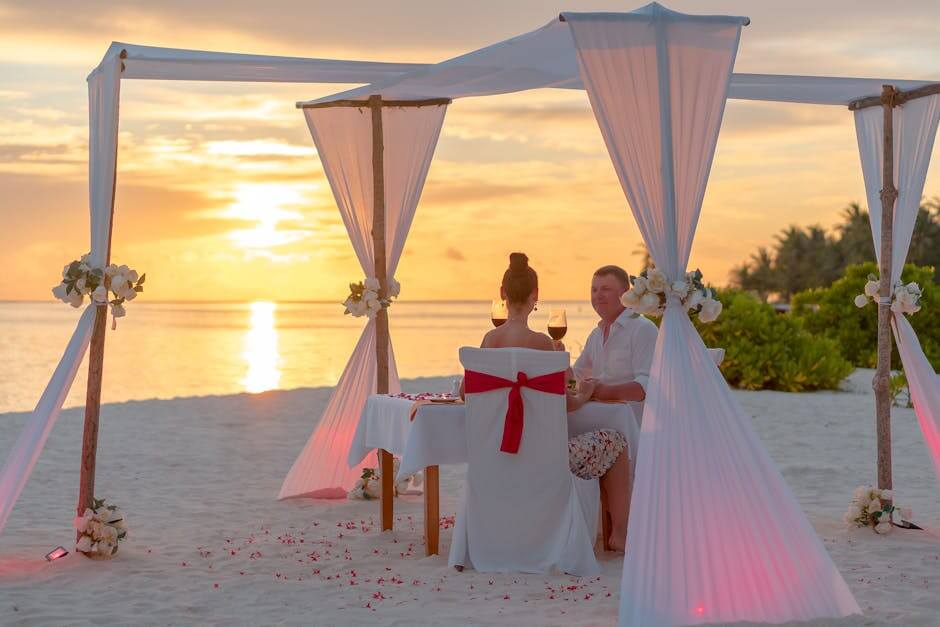 A couple enjoys a romantic dinner on a beach under a canopy at sunset, surrounded by a serene ocean view.