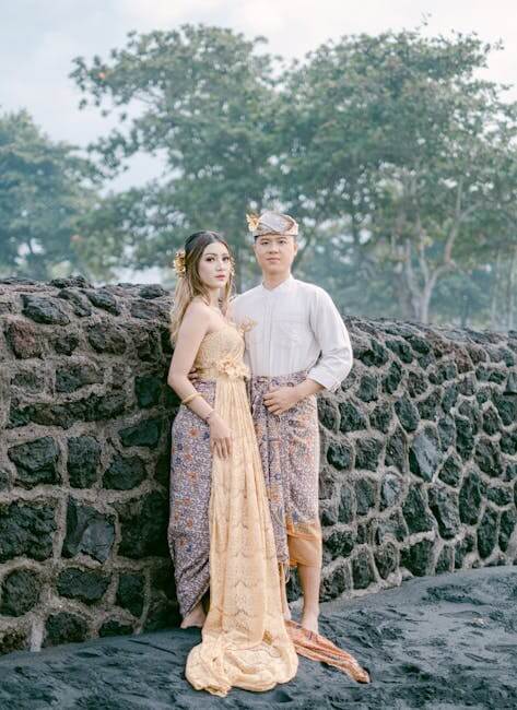 Couple in traditional Balinese attire poses next to stone wall outdoors.