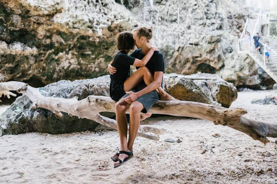 A couple shares an intimate moment on driftwood at a scenic beach location.