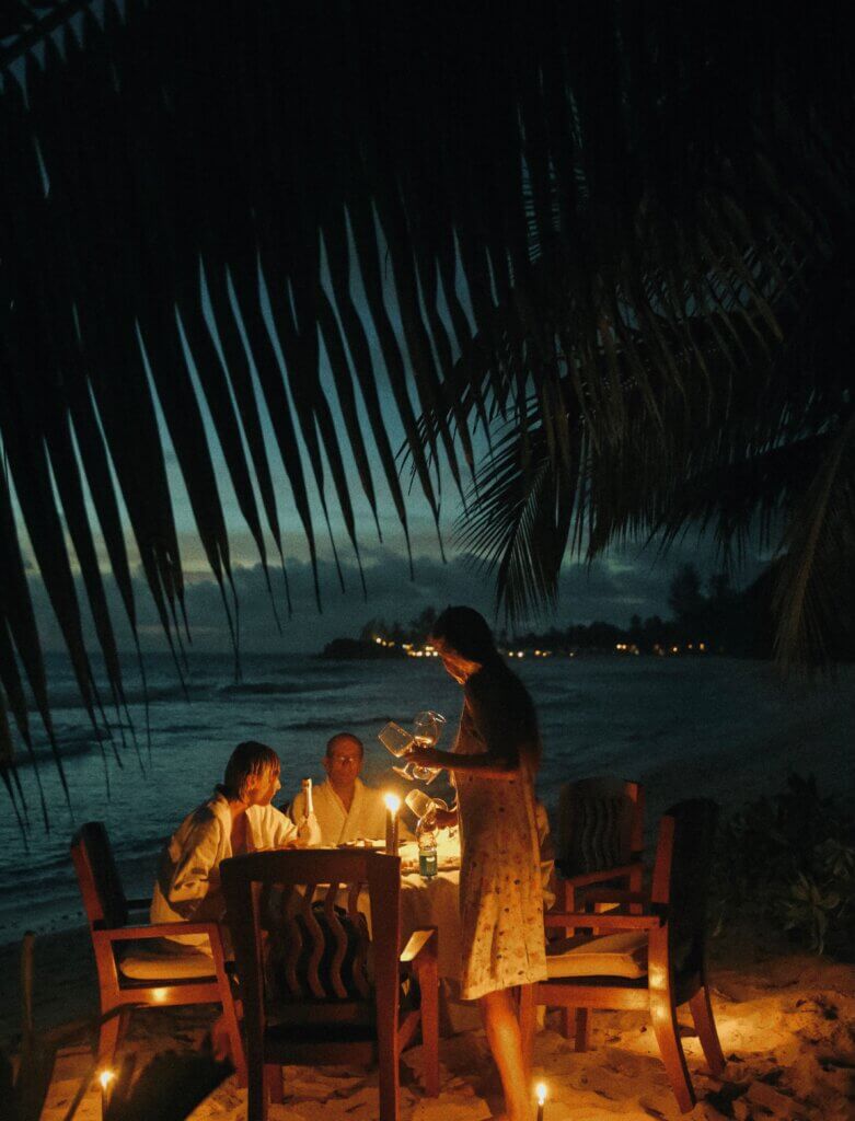 A serene beachside dining experience with friends illuminated by candlelight under palm trees.