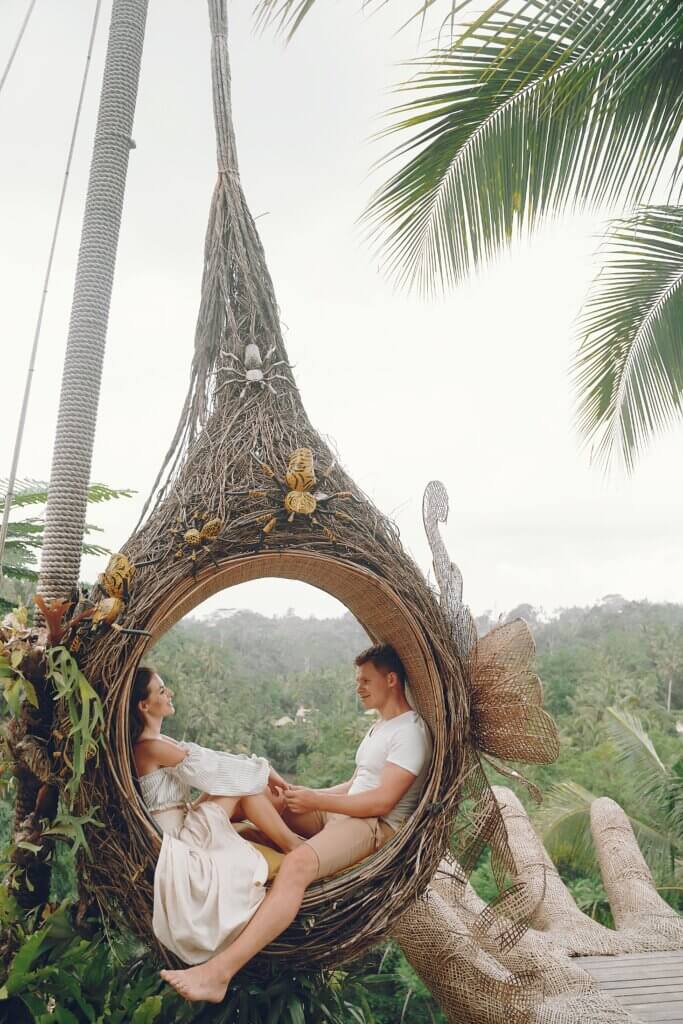 Side view full length of happy barefoot couple in white clothes holding hands and looking at each other while resting in big wicker hanging swing in tropical countryside in daytime
